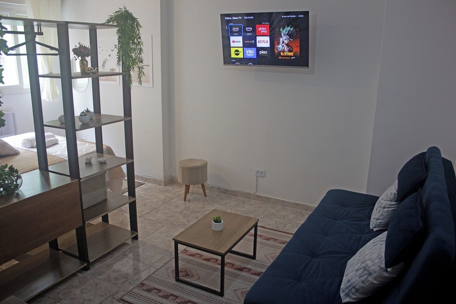 A living room with a sofa, a coffee table, a TV mounted on the wall displaying streaming services, and a shelving unit with decorative items.