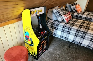 Ein Pac-Man-Spielautomat neben einem Bett mit Karo-Muster und Kissen.