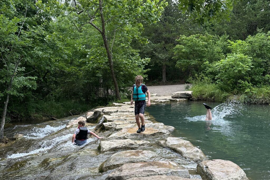 Un paysage naturel avec un ruisseau rocheux, des arbres et une personne debout sur des rochers.