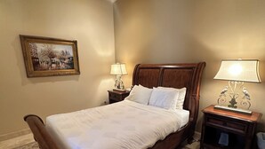 Een slaapkamer met een bed, een plafondventilator, een nachtkastje met een lamp, een ingelijst kunstwerk aan de muur en een kamerbreed tapijt met een patroon.