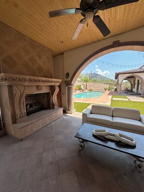 An outdoor patio with a stone fireplace, a ceiling fan, a sofa, and a view of a swimming pool and desert landscape. - Guest bedroom located outside luxury home (Tucson)