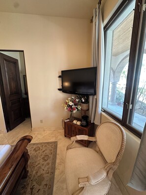 A room with a television mounted on the wall, a wooden side table with a vase of flowers, a wicker chair, and a large window with curtains. - Guest bedroom located outside luxury home (Tucson)
