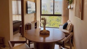 A modern dining area with a round table and wooden chairs, a TV mounted on the wall, and a view of buildings through large windows. - 10 min. from beach, New luxury condo w/ Pool, Gym, Sauna, Parking and laundry! (Playa del Carmen)