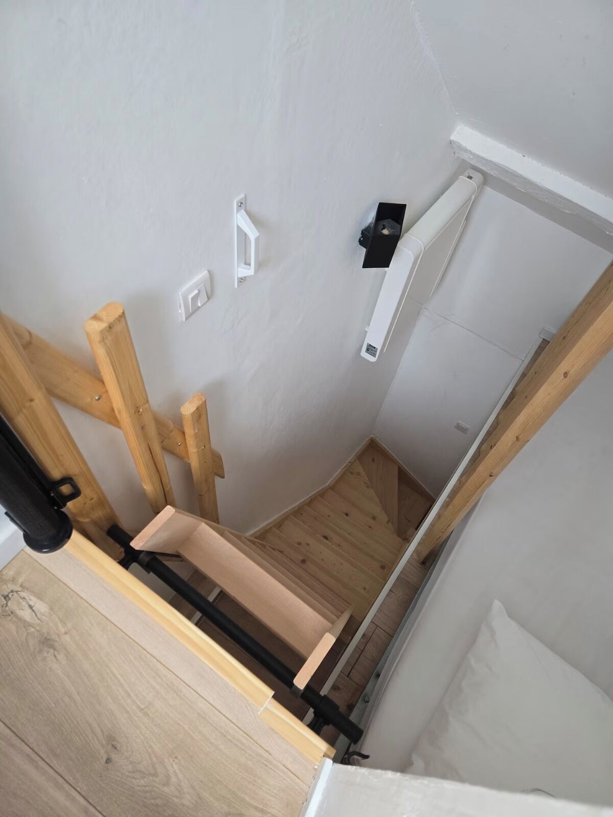 A narrow, wooden staircase with a handrail, white walls, and a small black shelf.