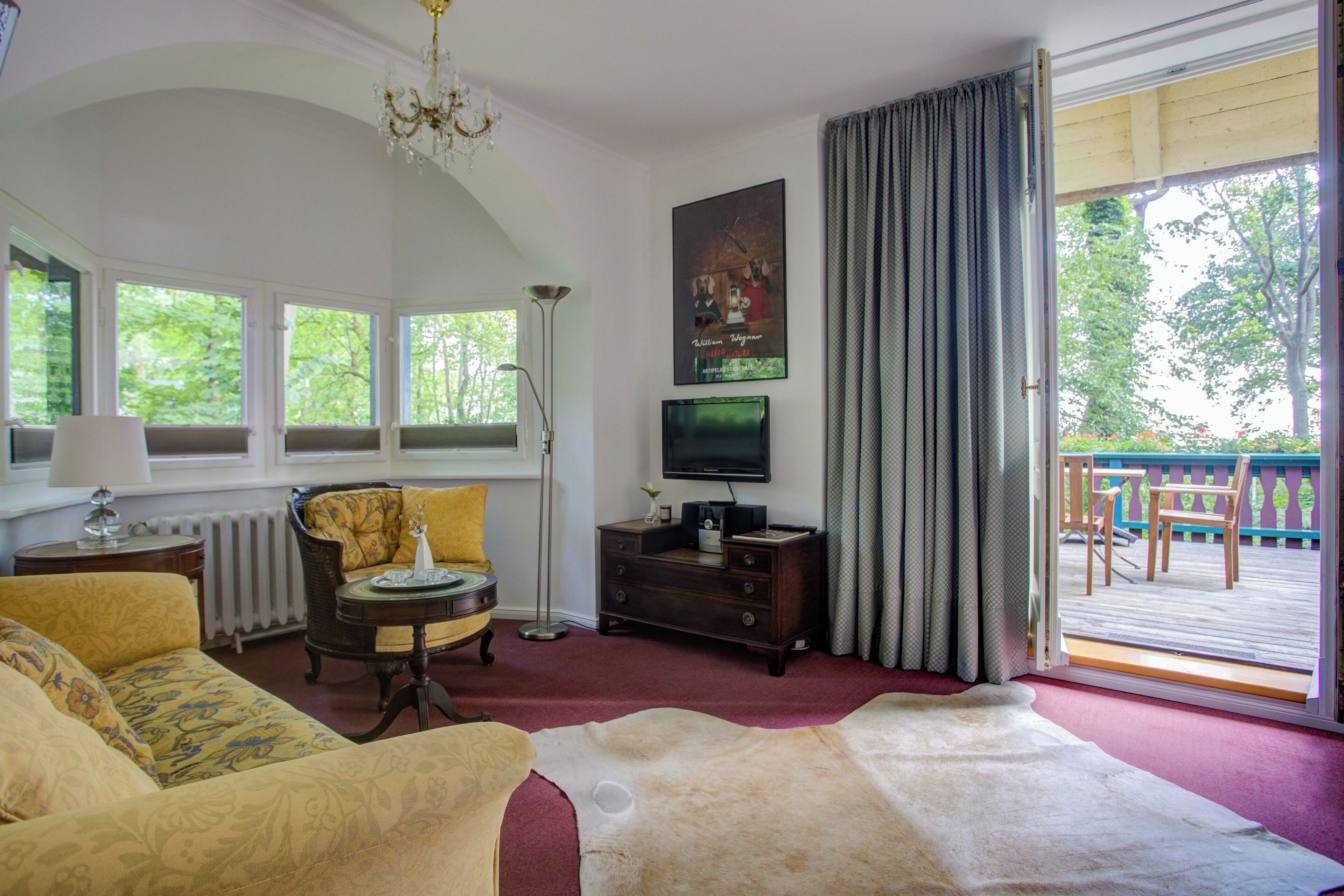 Un salon avec un canapé, un fauteuil et un grand tapis. On y trouve un téléviseur posé sur un meuble et une fenêtre avec des rideaux.