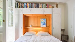 A bedroom with a wooden headboard, a bed with white bedding, a bookshelf above the bed, and a small table with a chair. - Strandvilla Hubertus - H1 - Marc Chagall (Ostseebad Boltenhagen)