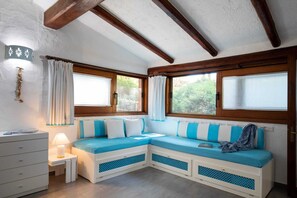 Ein gemütliches Zimmer mit einer Eckcouch, einem kleinen Nachttisch und einer Lampe. Zwei Fenster mit Vorhängen, und die Zimmerdecke ist aus rustikalem Holz.