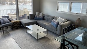 A modern living room with a grey sectional sofa, a glass coffee table, and a view of a body of water through large windows.