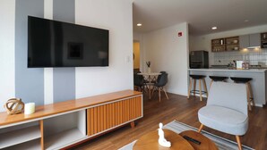 A modern living room with a flat-screen TV mounted on the wall, a wooden entertainment unit, a grey armchair, and a dining area with a round table and chairs. - w* | Chic & Inviting 2BR w/ Balcony in Miraflores (Miraflores)