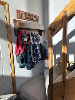 A wooden coat rack with jackets and a plaid shirt hanging on it, a wooden bench, and a welcome sign above the coat rack. - Green Mountain Getaway (Munds Park)