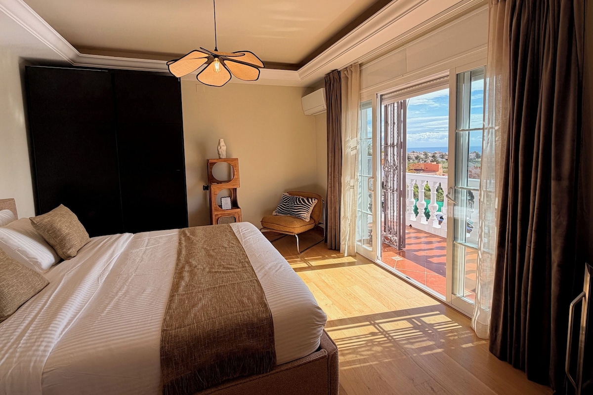 A hotel room with a large bed, a wooden nightstand, a chair, and a balcony with a view.