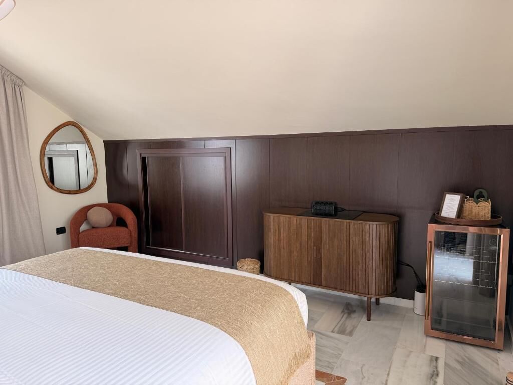 A hotel room with a large bed, a wardrobe, a mirror, a small table, and a minibar.