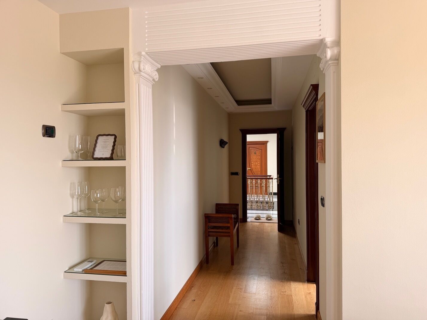 Un couloir aux murs blancs, doté d’un plancher en bois et comprenant une chaise en bois. On y trouve des étagères avec des verres à vin et un certificat encadré.