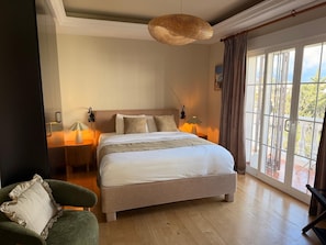 Ein Schlafzimmer mit einem großen Bett, Nachttischlampen, einem Sessel und Blick durch ein Fenster.
