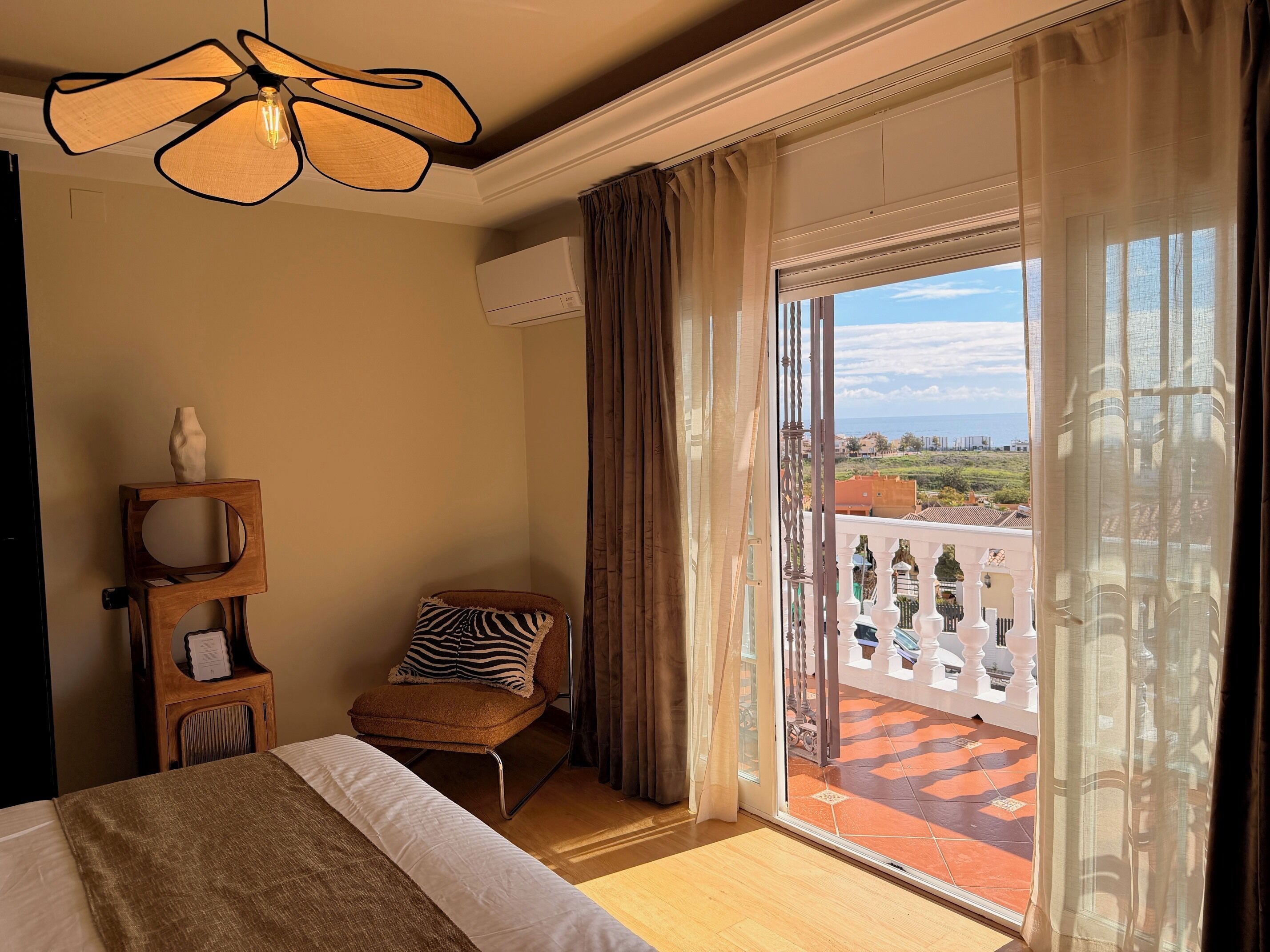 Ein Hotelzimmer mit einem Bett, einem Sessel, einem kleinen Tisch mit einer Vase, einem Deckenventilator und einem großen Fenster mit Blick auf die Stadt.