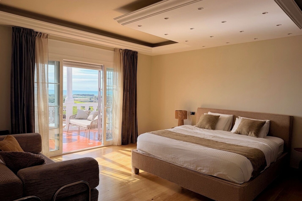 A hotel room with a large bed, a balcony with a view, and a seating area with chairs.