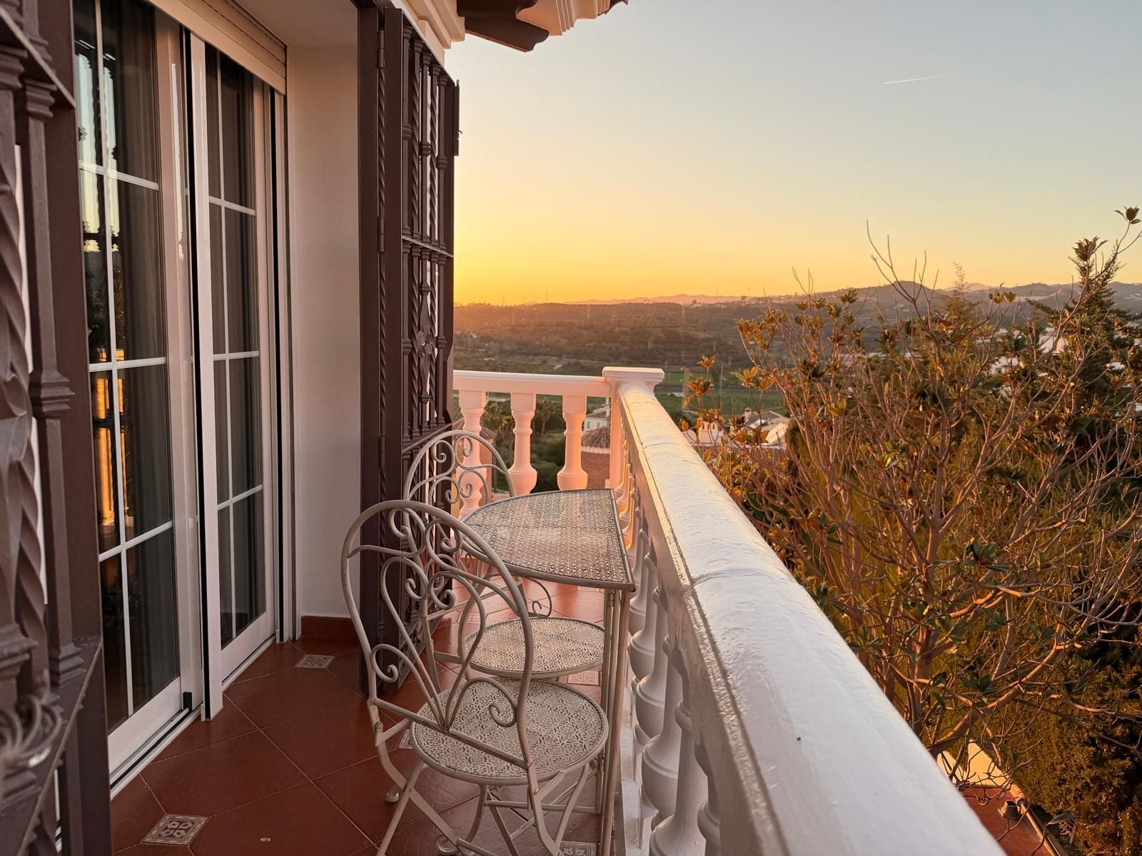 A balcony with two chairs and a table, overlooking a scenic view with mountains and vegetation.