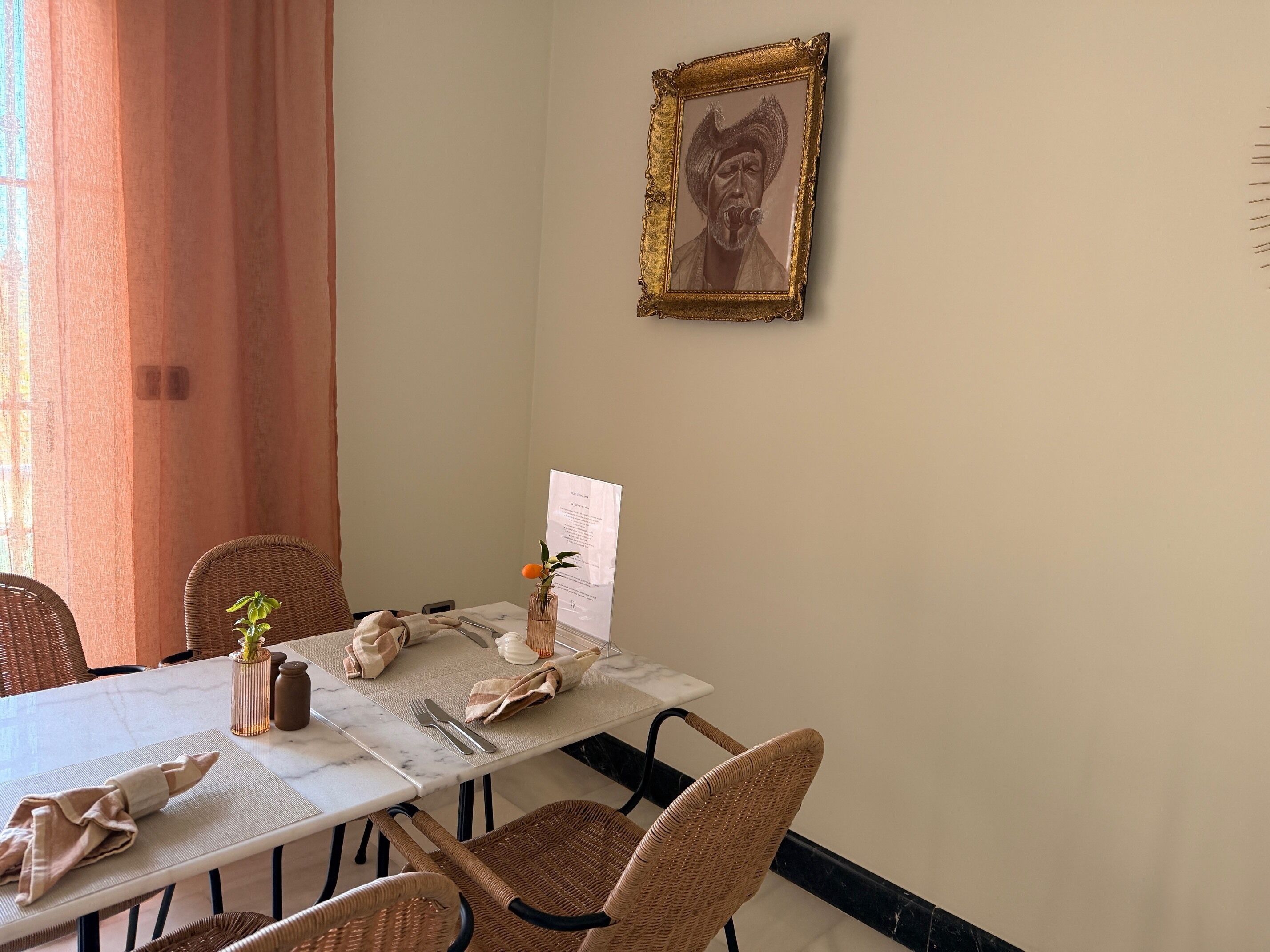 Een eetruimte met een witte tafel gedekt voor twee personen, rieten stoelen, een vaas met een plant en een ingelijst beeld aan de muur.