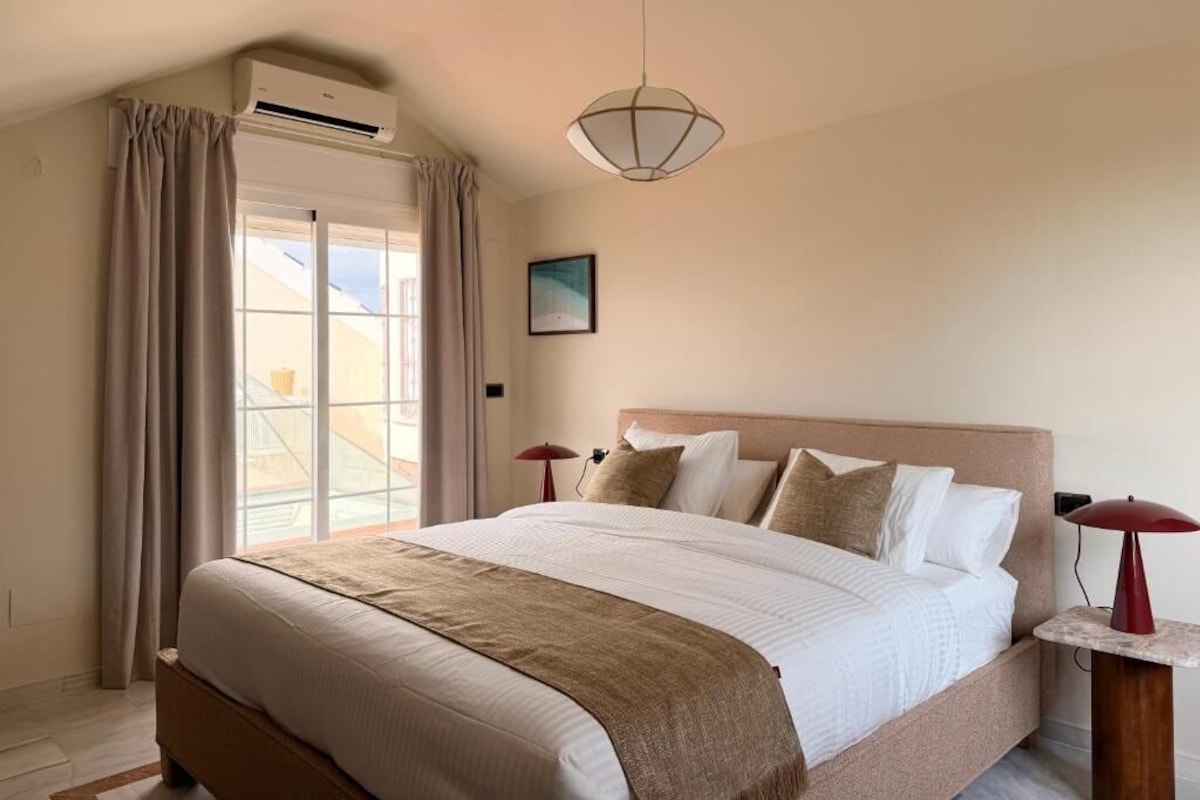 A bedroom with a large bed, an air conditioning unit, a hanging light fixture, and a view of the outside through a window with curtains.