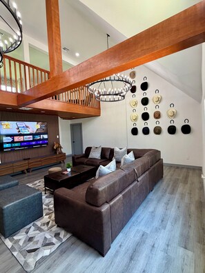 Ein modernes Wohnzimmer mit einem braunen Ledersofa, einer Holztreppe, einer Chandelüster und einem an der Wand befestigten Fernseher.