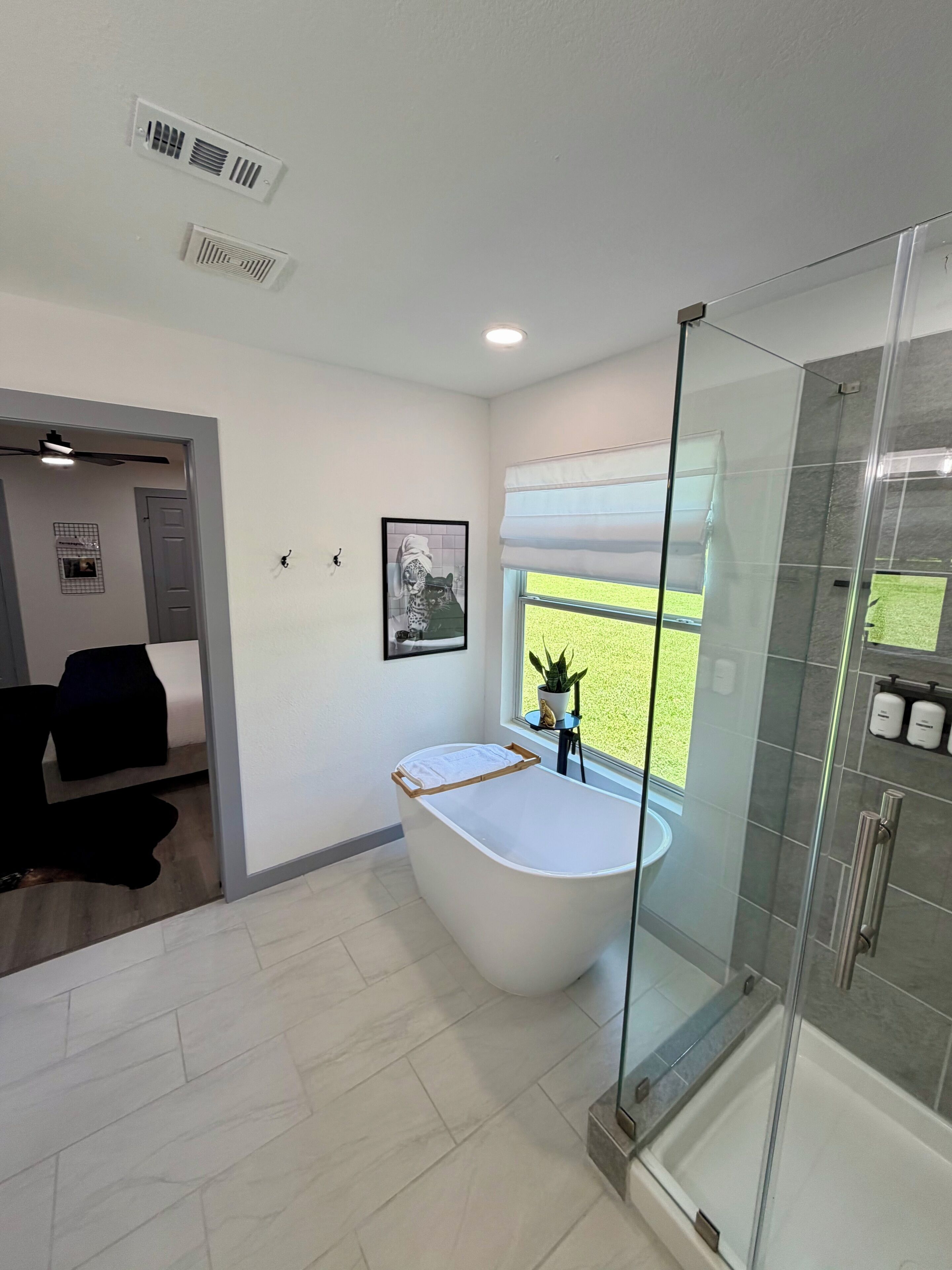 Un baño moderno con ducha envuelta en vidrio, una bañera independiente y un ventanal con vistas a un césped verde.