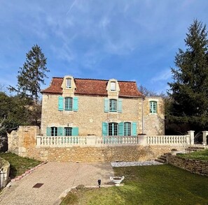 A stone building with a red-tiled roof, multiple windows with green shutters, and a stone fence.