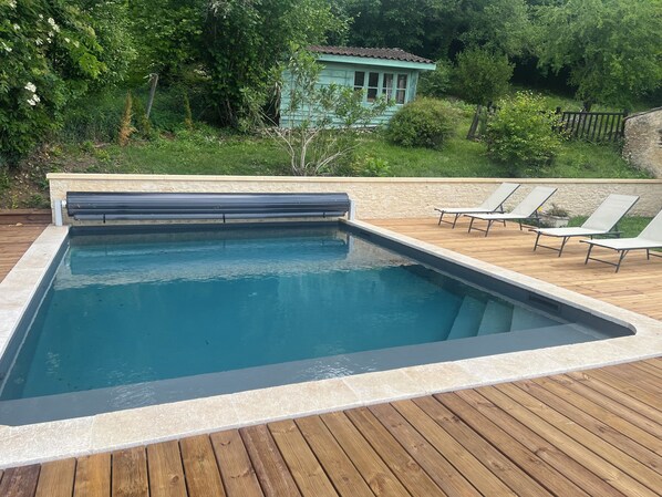 A swimming pool with a wooden deck, surrounded by greenery and a small building.