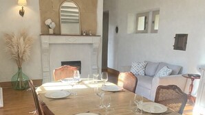 Uma sala de jantar com uma mesa de madeira posta para seis pessoas, rodeada por cadeiras de vime. É visível uma lareira com um espelho acima, e um candelabro pendurado no teto.