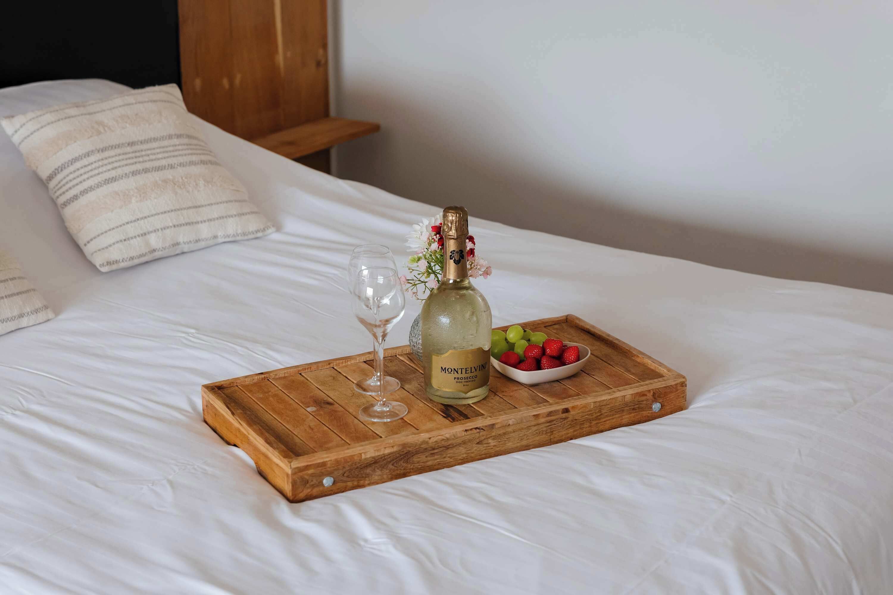 A bed with a wooden tray holding a bottle of Montelvino Prosecco DOCG, a glass of wine, and a bowl of grapes and strawberries.