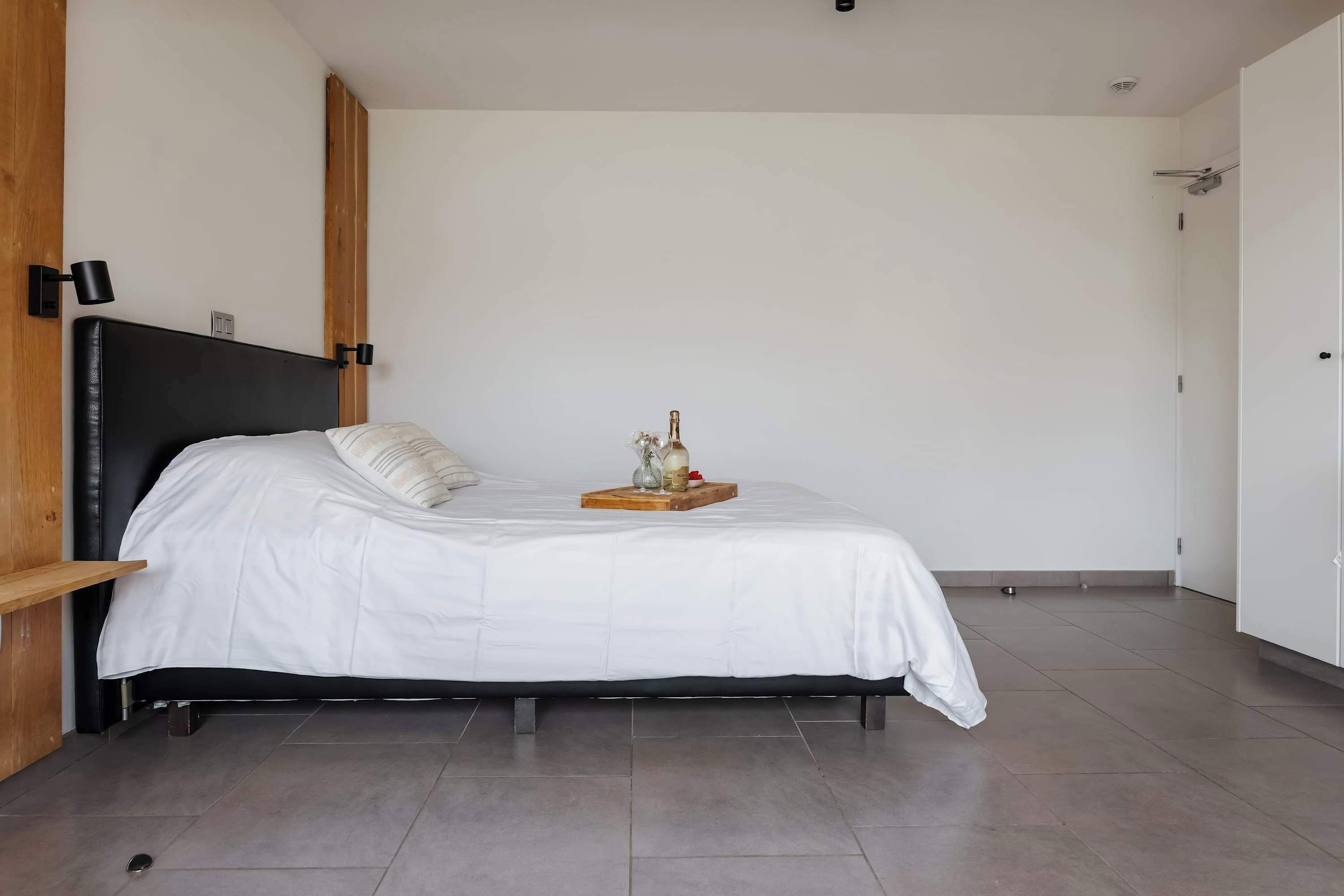 A bedroom with a bed, a bedside table, a nightstand, a wall-mounted lamp, and a door.
