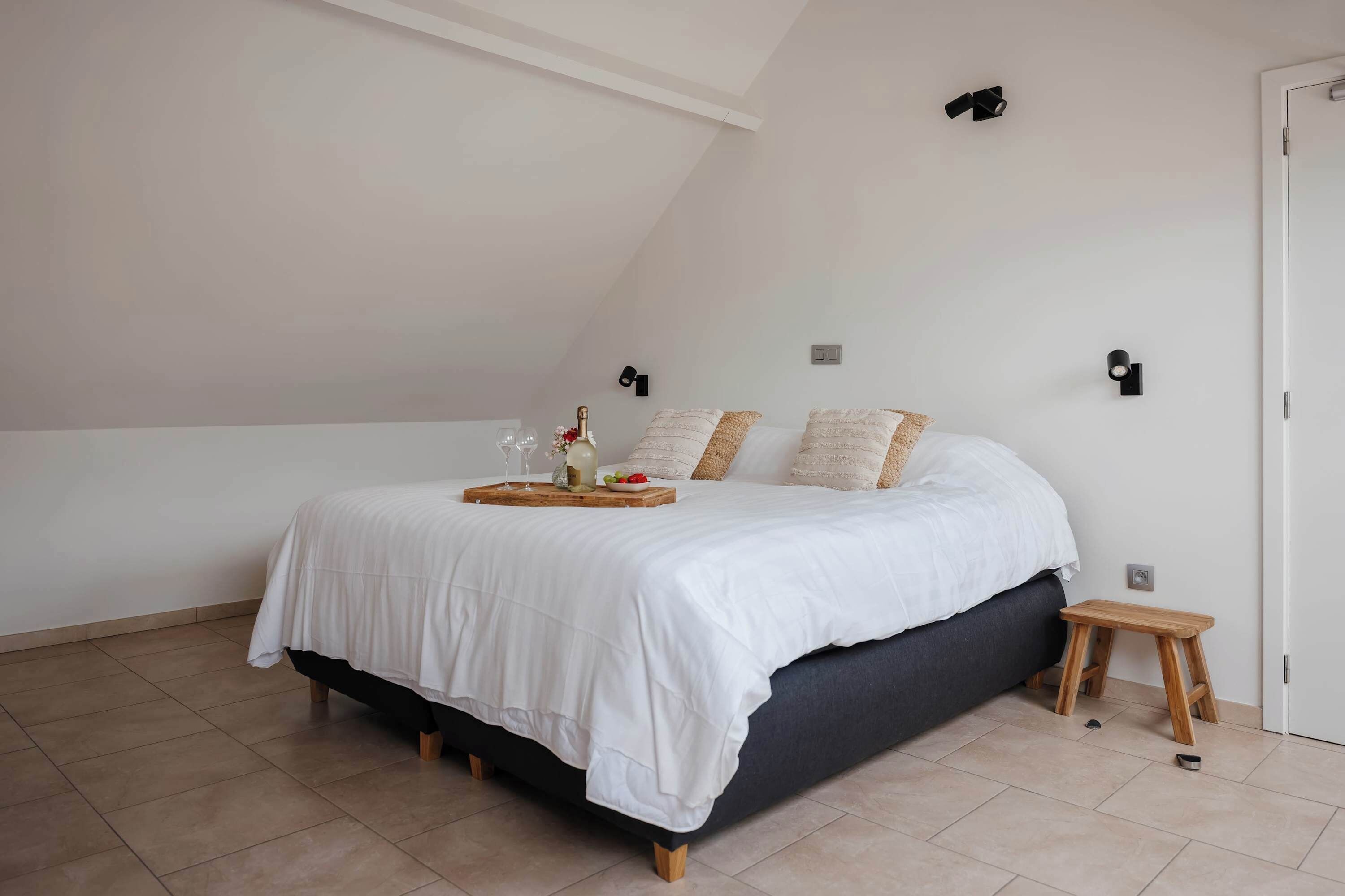 A bedroom with a bed, a small wooden stool, and a nightstand with a tray.