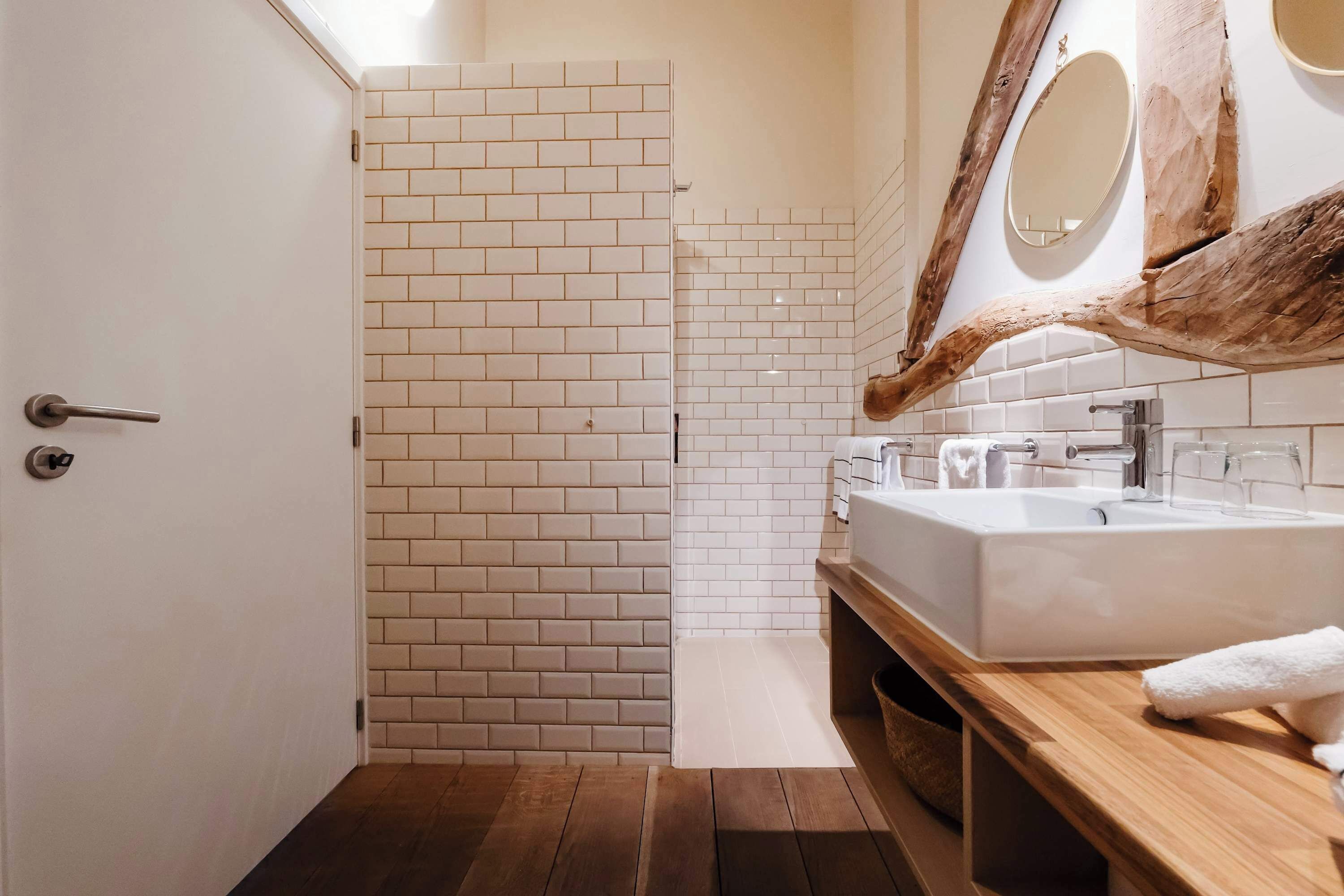 Un baño con puerta blanca, ducha con azulejos blancos, tocador de madera con lavabo y espejo con marco de madera.