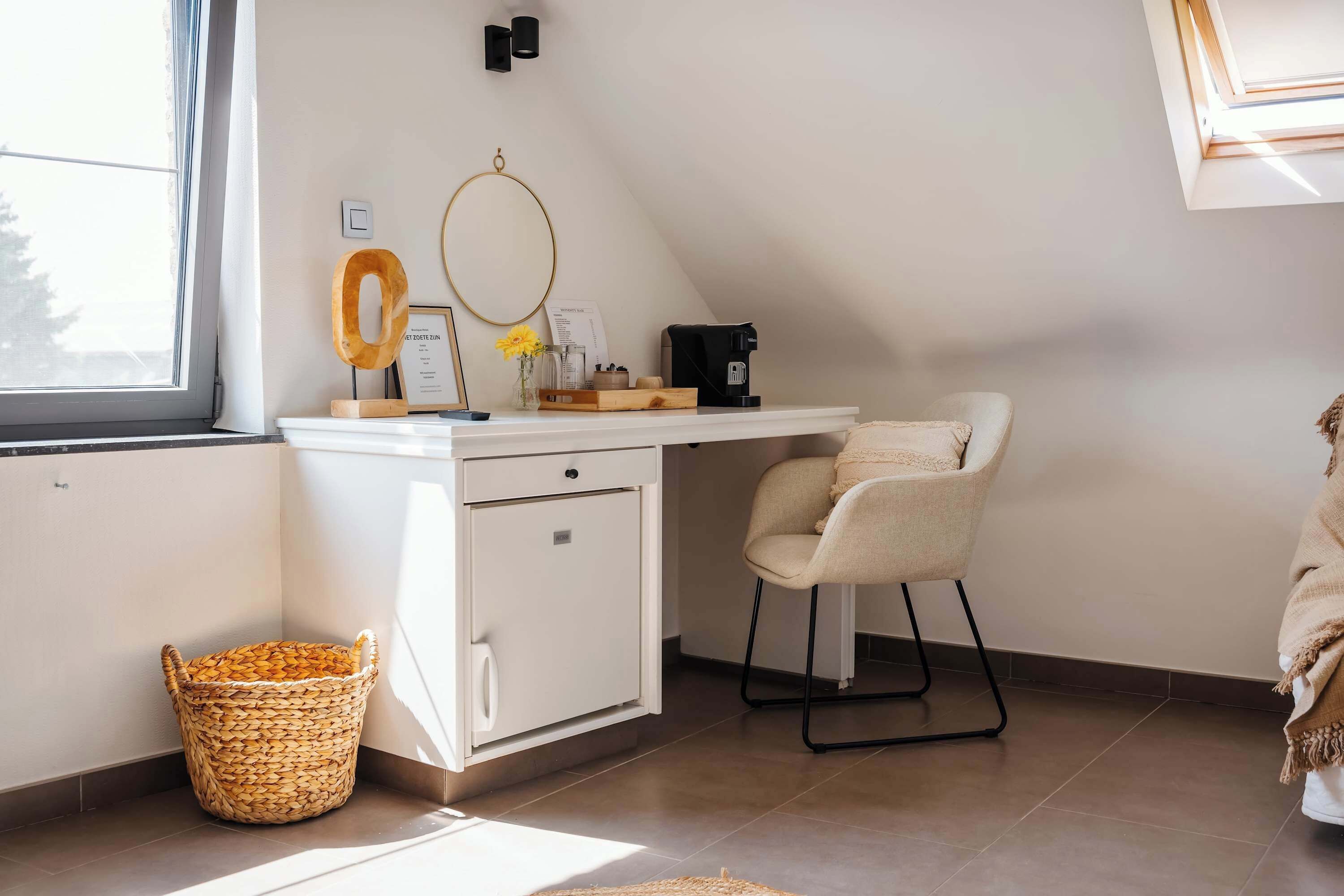 Une pièce avec un bureau blanc, une chaise, un petit meuble, un panier en osier et une fenêtre.