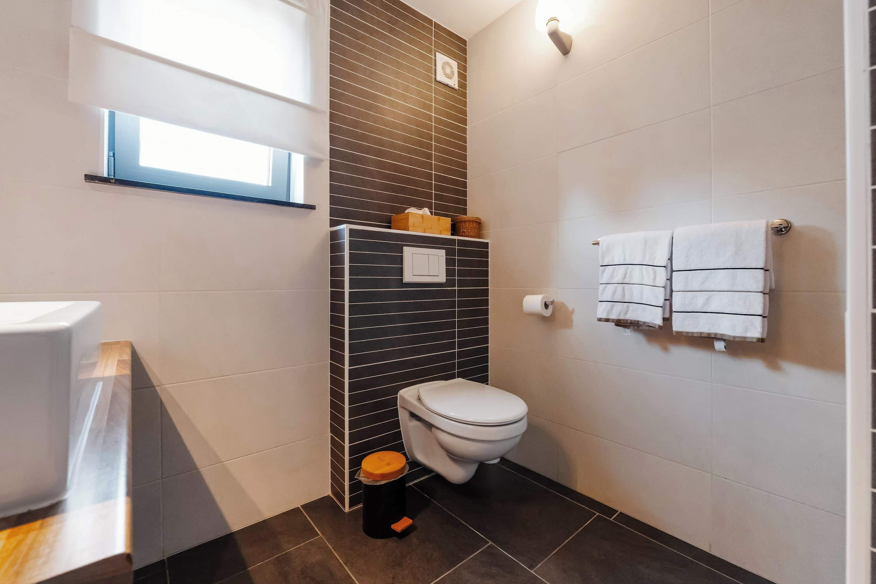 A modern bathroom with a toilet, a trash bin, a towel rack, and a window.