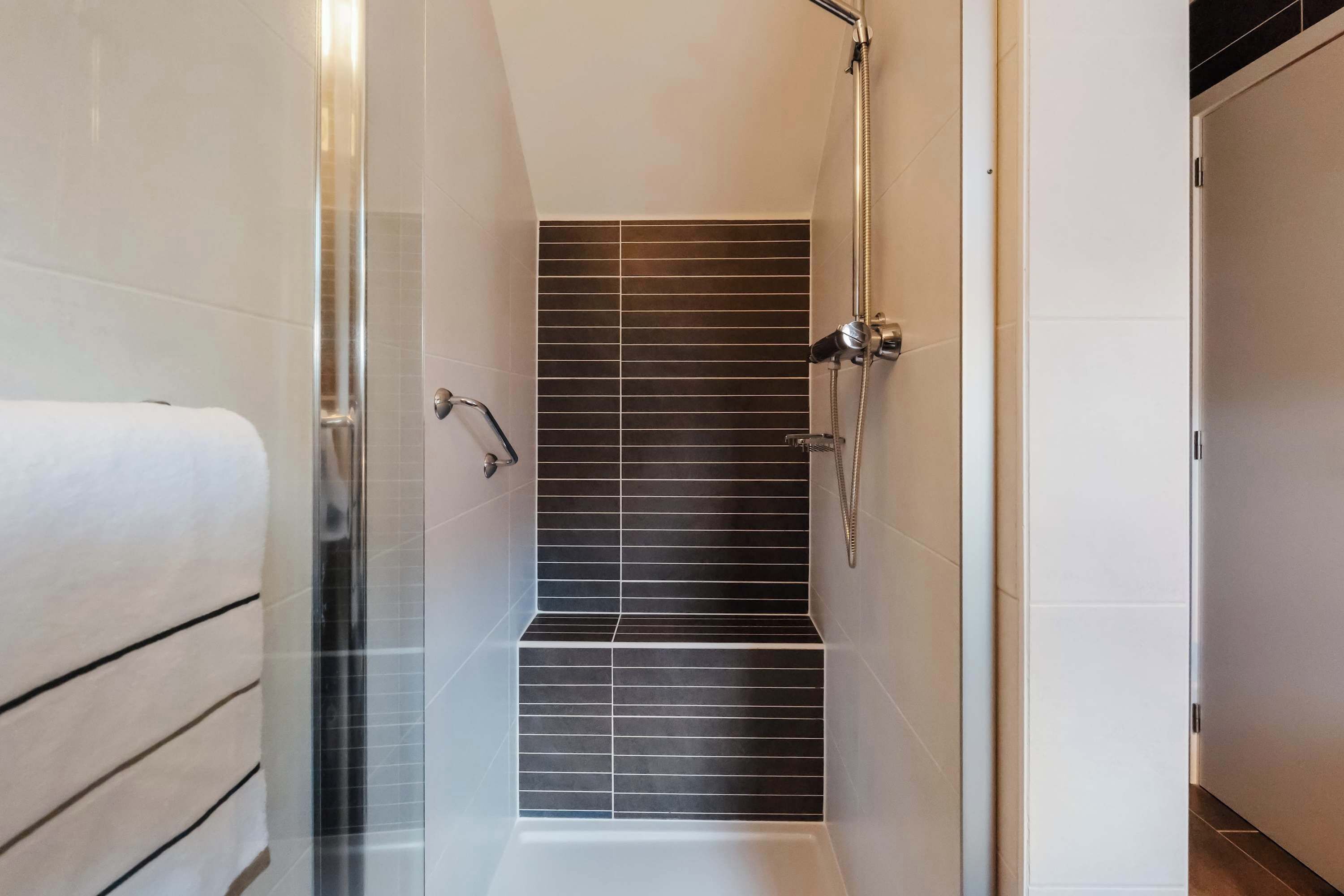 Une salle de bain moderne avec une douche à l’italienne en verre, une serviette blanche et des murs recouverts de carreaux foncés.