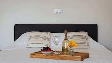 Una cama bien hecha con un cabecero oscuro, dos almohadas, una bandeja con una botella, un vaso y un cuenco de fruta.