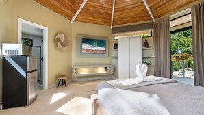 Ein modernes Schlafzimmer mit Holzboden, einem Bett mit weißer Bettwäsche, einem Fernseher, einem kleinen Hocker und Blick ins Grüne.