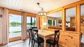 A wooden interior with a dining table and chairs, a sofa, and large windows offering a view of a lake. - Stunning Lakefront Cottage with Beach & Hot Tub (Alger)