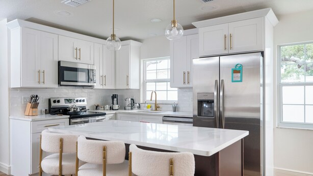 Una cocina moderna con armarios blancos, electrodomésticos de acero inoxidable, una isla central con taburetes y luces colgantes.