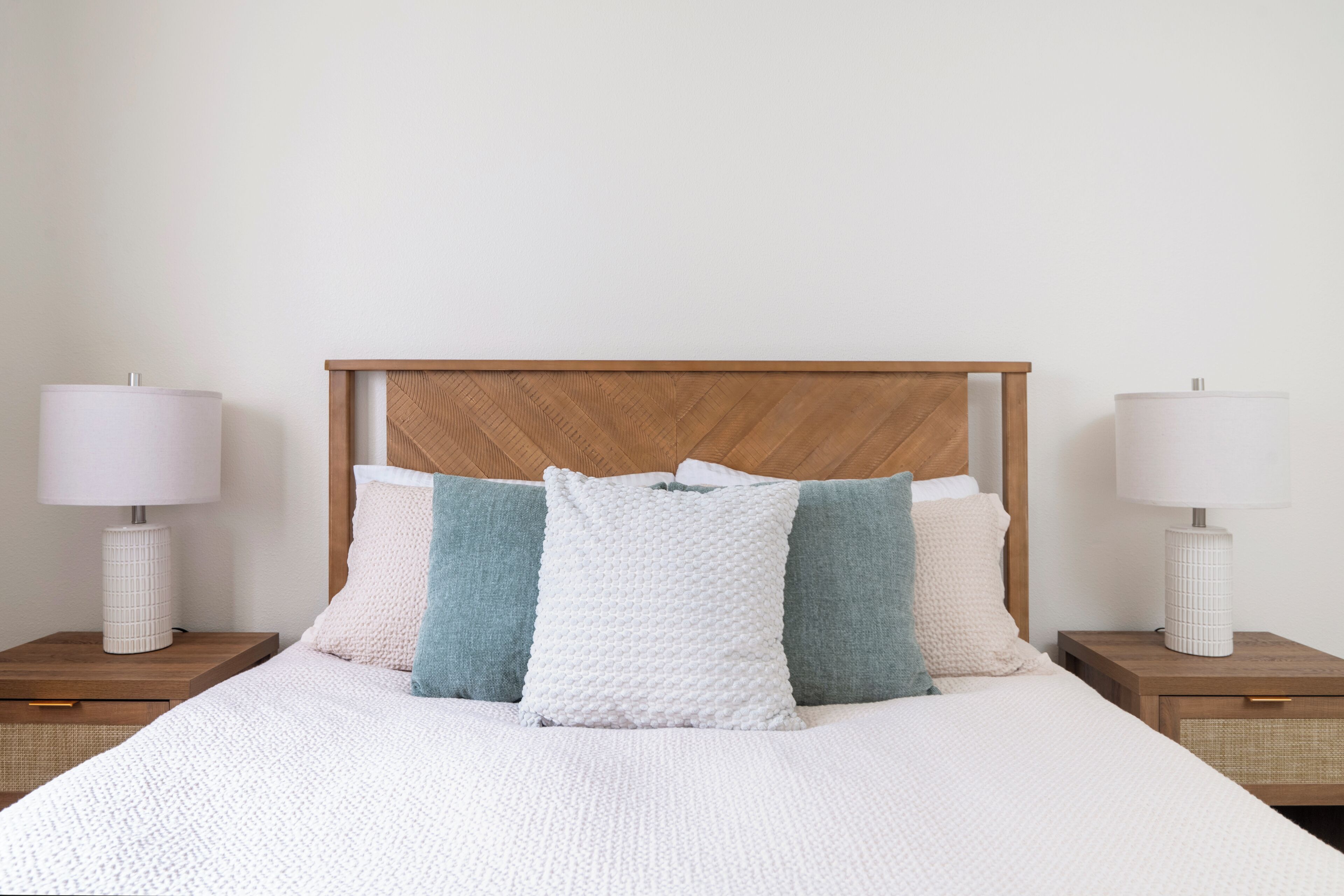 Ein ordentlich bezogenes Bett mit Holz-Kopfteil, zwei Nachttische mit Lampen und eine schlichte Wand.