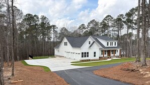 A two-story house with a driveway leading to a garage, surrounded by a wooded area. - Masters Rental - Brand New Build (Evans)