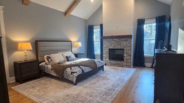 A bedroom with a sloped ceiling, a fireplace, a bed with patterned bedding, a desk, and a large window with curtains. - Masters Rental - Brand New Build (Evans)