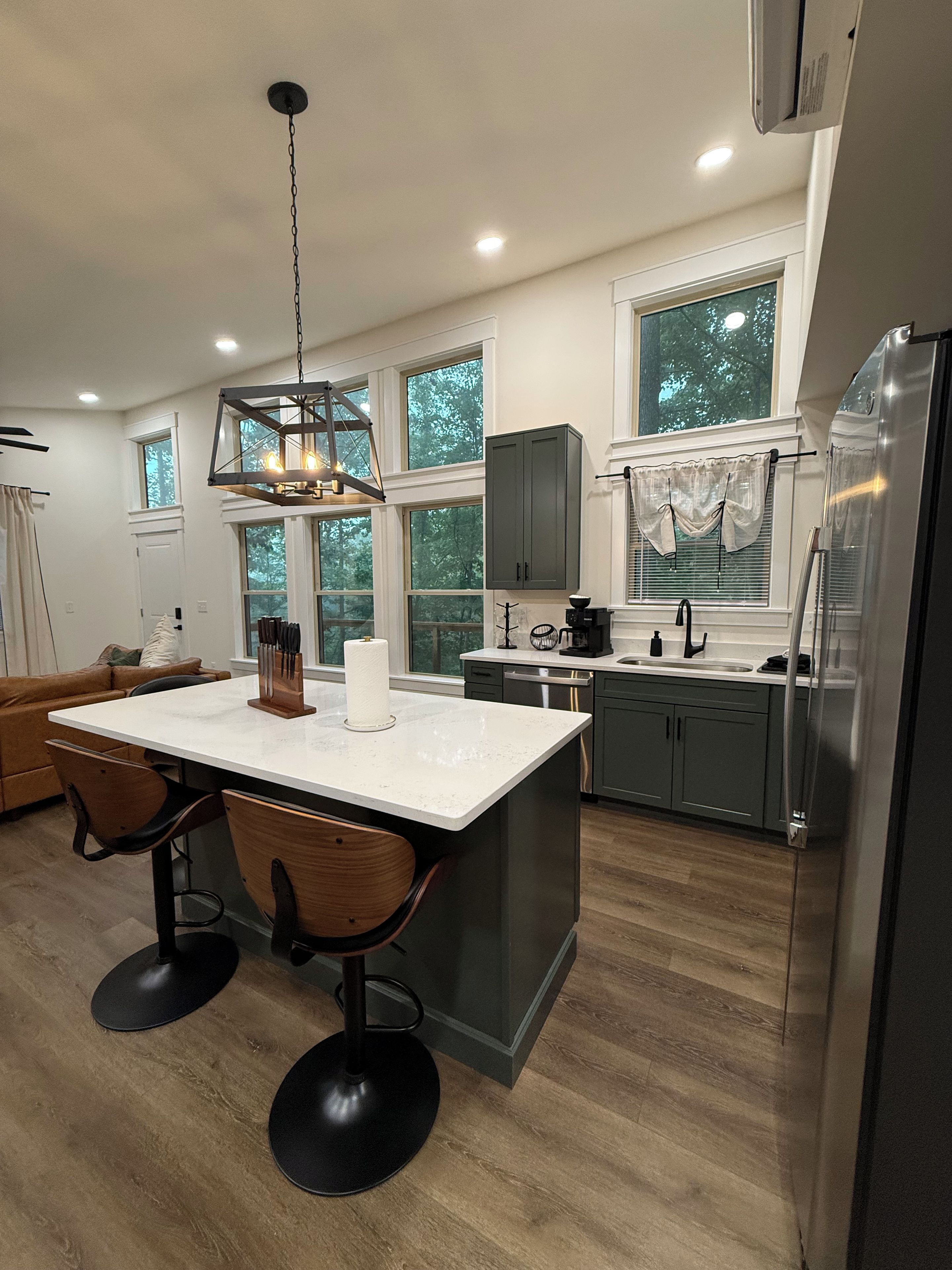 A modern kitchen with a central island, wooden chairs, a hanging light fixture, and a large window.