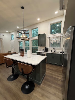 A modern kitchen with a central island, wooden chairs, a hanging light fixture, and a large window.