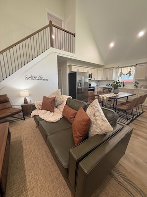 A spacious living area with a high ceiling, a staircase, a kitchen, and a dining area.