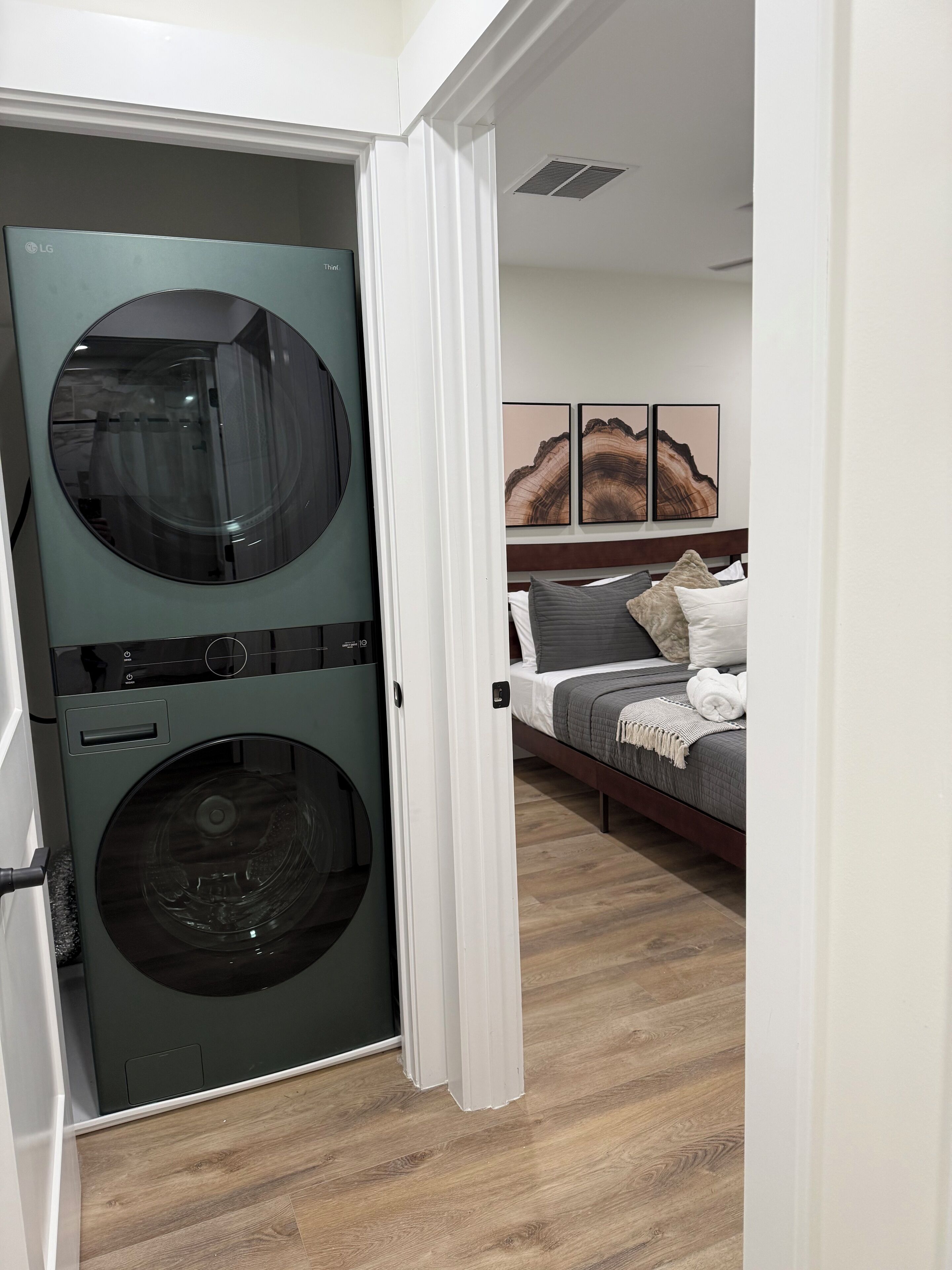 A laundry room with a front-loading washer and dryer, a bed with a gray and white bedspread, and a wall with three framed artworks.