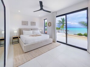 Ein modernes Schlafzimmer mit einem großen Bett, einem Deckenventilator und Blick auf Strand und Meer durch eine Schiebetür.