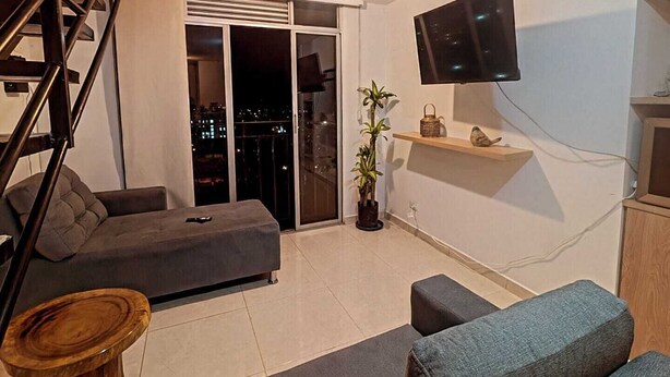 Un salon moderne avec un canapé gris, une table basse en bois, un téléviseur fixé au mur et un balcon offrant une vue sur la ville.