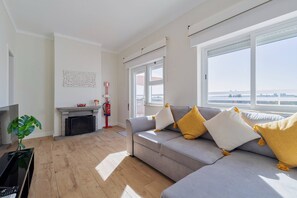 A modern living room with a grey sofa, a flat-screen TV, a fireplace, and a large window offering a view of the city. - Serra&Rio no Sado - By Y Concept (Setúbal)