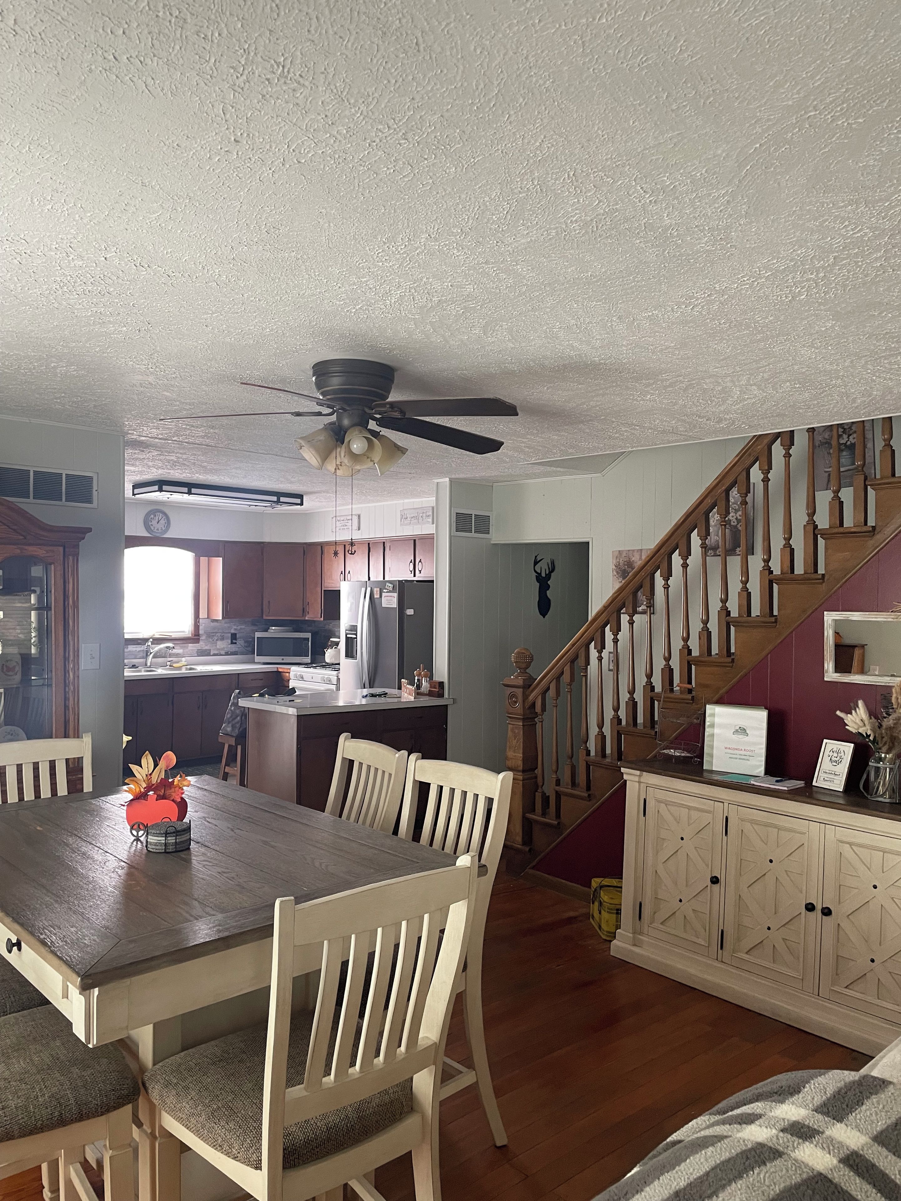 A dining area with a wooden table and chairs, a kitchen in the background, and a staircase leading to another level.